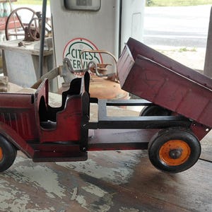 May include: A vintage red and black toy dump truck with orange wheels. The truck bed is raised, showcasing its hauling capability. The toy truck is made of metal and has a classic design.