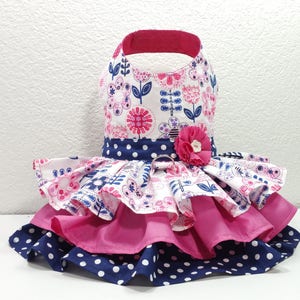 May include: A dog dress with a floral print bodice and a layered skirt. The bodice has a white background with pink and blue floral patterns. The skirt has layers of pink and navy blue polka dot fabric. A pink flower adorns the waist.