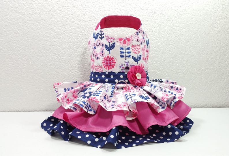 May include: A dog dress with a floral print bodice and a layered skirt. The bodice has a white background with pink and blue floral patterns. The skirt has layers of pink and navy blue polka dot fabric. A pink flower adorns the waist.