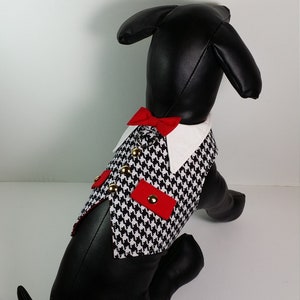 Puede incluir: Un chaleco para perro de pata de gallo blanco y negro con detalles rojos y botones dorados. El chaleco tiene un cuello blanco y una pajarita roja.