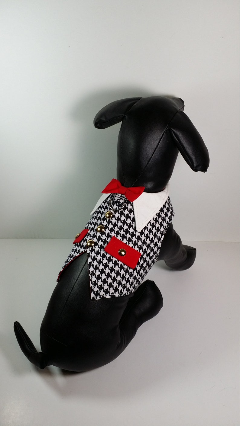 Puede incluir: Un chaleco para perro de pata de gallo blanco y negro con detalles rojos y botones dorados. El chaleco tiene un cuello blanco y una pajarita roja.
