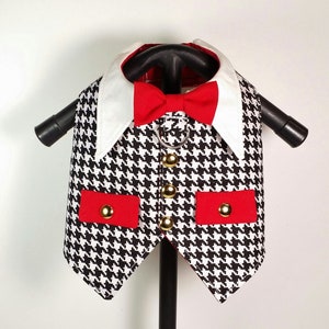 Puede incluir: Un chaleco para perro de color negro y blanco con estampado de pata de gallo, con una pajarita roja y botones dorados. El chaleco tiene dos bolsillos rojos con botones dorados.