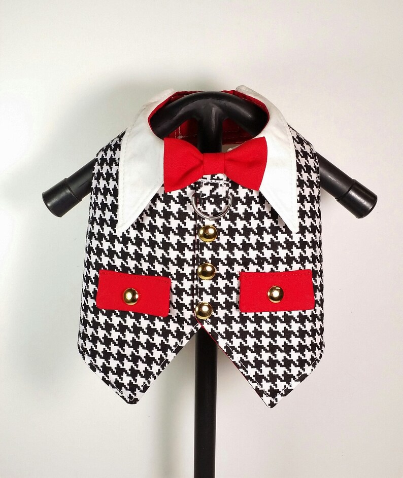 Puede incluir: Un chaleco para perro de color negro y blanco con estampado de pata de gallo, con una pajarita roja y botones dorados. El chaleco tiene dos bolsillos rojos con botones dorados.