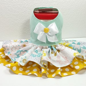 May include: A mint green dog dress with a white satin bow and yellow button detail. The skirt features layers of floral and polka dot fabric in yellow, pink, and blue. This pet apparel is designed to fit around a jar.