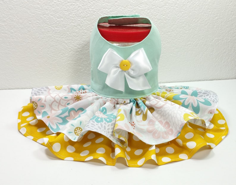 May include: A mint green dog dress with a white satin bow and yellow button detail. The skirt features layers of floral and polka dot fabric in yellow, pink, and blue. This pet apparel is designed to fit around a jar.