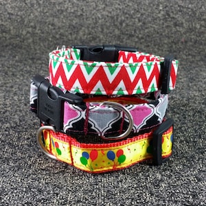 May include: Three dog collars stacked on top of each other. The top collar is red and green with a chevron pattern. The middle collar is black and gray with a pink trim and a geometric pattern. The bottom collar is yellow with red and blue balloons and confetti.