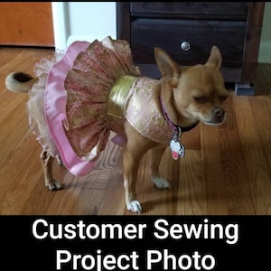 May include: A small brown dog wearing a pink and gold dress. The dress features a ruffled skirt and a gold band around the waist. The dog is standing on a wooden floor. Text at the bottom reads "Customer Sewing Project Photo."