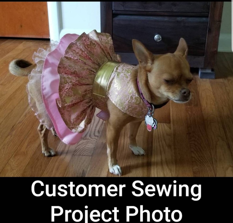 May include: A small brown dog wearing a pink and gold dress. The dress features a ruffled skirt and a gold band around the waist. The dog is standing on a wooden floor. Text at the bottom reads "Customer Sewing Project Photo."