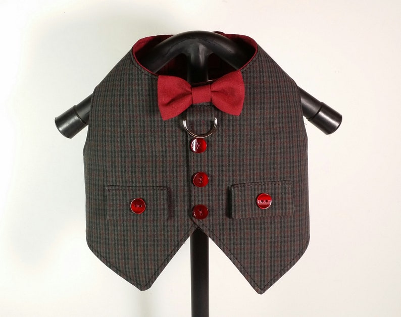 Puede incluir: Un chaleco para perro de cuadros grises y rojos con una pajarita roja y botones rojos. El chaleco tiene dos bolsillos y un anillo de metal para sujetar una correa.