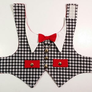 Puede incluir: Un chaleco para perro de pata de gallo negro y blanco con detalles rojos y un cuello blanco con una pajarita roja. El chaleco tiene dos bolsillos rojos con botones dorados y correas ajustables.