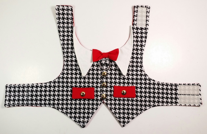 Puede incluir: Un chaleco para perro de pata de gallo negro y blanco con detalles rojos y un cuello blanco con una pajarita roja. El chaleco tiene dos bolsillos rojos con botones dorados y correas ajustables.