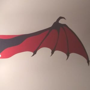 Könnte beinhalten: Ein flacher, gestanzter Aufkleber aus Papier oder Vinyl in Form eines einzelnen Fledermausflügels. Der Flügel hat eine tiefrote Membran mit schwarzen Rippen und einen weißen, dekorativen, gewellten Befestigungspunkt auf der linken Seite.