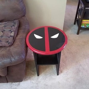 May include: A small, round side table with a black base and a tabletop featuring a Deadpool design. The tabletop has a black background with a red border and a red vertical stripe. White eye shapes complete the design.