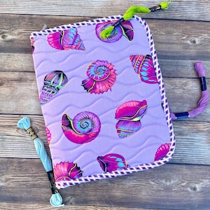 May include: A quilted fabric case in light purple featuring a repeating pattern of vibrant pink, blue, and purple seashells. The case has a white and purple checkered border and includes three separate embroidery floss skeins in lime, purple, and light blue.