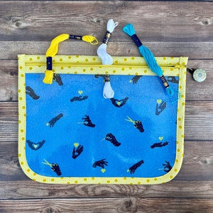 May include: A clear zippered pouch with a blue background and a pattern of black hands holding hearts. The pouch has a yellow border and a zipper pull with a blue and white floral design. Three spools of embroidery floss are visible, one yellow, one white, and one blue.