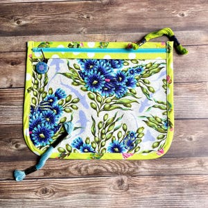 May include: A rectangular, zippered craft pouch with a floral design. The pouch features a white background with blue flowers and green leaves. The edges are outlined in a bright green, and a blue button is attached to the zipper.