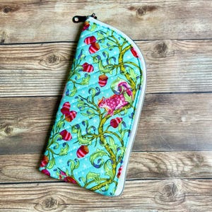 May include: A quilted, zippered pouch with a whimsical design. The teal fabric features a pattern of stylized trees, red acorns, and a pink squirrel. The pouch has a white zipper and a black pull.