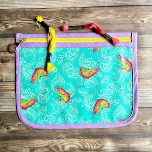 May include: A rectangular, zippered pouch with a teal background featuring a repeating pattern of colorful fish and white wave designs. The pouch has a yellow zipper, a purple border, and a decorative button. Two spools of thread are on top.