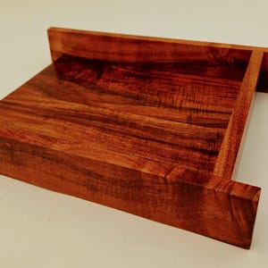May include: A handcrafted wooden tray with a rich, reddish-brown hue. The tray features a rectangular design with raised edges and a sloped interior, showcasing the natural wood grain. The tray's dimensions are approximately 12 inches long, 8 inches wide, and 2 inches high.