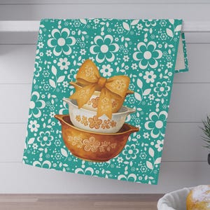 Może przedstawiać: Turkusowy ręcznik kuchenny z białym kwiatowym wzorem wisi na białej półce. Dwie miski w stylu vintage z kwiatowym wzorem są ułożone w stos, zwieńczone złotą kokardą. Mała roślina w czarnej doniczce i kosz z chlebem znajdują się na pierwszym planie.