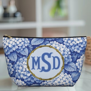 Puede incluir: Un estuche con cremallera que presenta un estampado floral de hortensias azules y blancas. En el centro hay un emblema circular con un borde estilo bambú, que contiene las letras azules del monograma MSD sobre un fondo azul claro.
