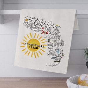 Puede incluir: Un paño de cocina blanco con un mapa de Florida, con la palabra "Florida" en cursiva y la ciudad de Jacksonville con el código postal 32258. El mapa incluye ilustraciones de un sol y varias ciudades de Florida.