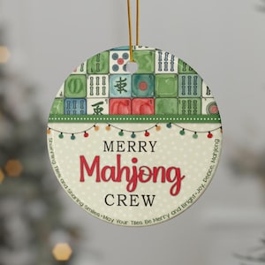 May include: Circular Christmas tree decoration with a green and cream design. The ornament features Mahjong tiles and the words "MERRY Mahjong CREW" in red. A string of lights and text are around the edge. It hangs from a gold string.