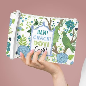 May include: White clutch with a floral and bird pattern, featuring blue and green accents. The design includes the text "BAM! CRACK! DOT! Cash!" in a decorative frame. The clutch has a wrist strap and a zipper closure.