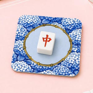 Könnte beinhalten: Ein quadratischer Untersetzer mit blauem Blumenmuster und einem weißen Mahjong-Stein mit einem roten chinesischen Schriftzeichen. Mehrere Mahjong-Steine mit blauen und weißen Details sind um den Untersetzer auf einer rosa Oberfläche angeordnet. Ein dekorativer Artikel für Spieleliebhaber.