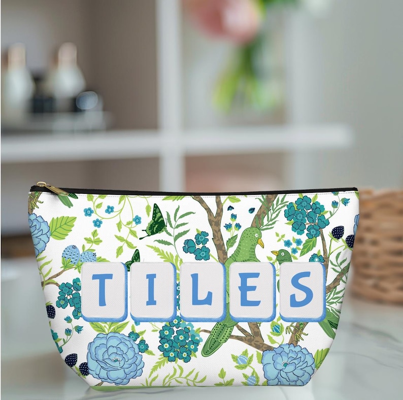 May include: White cosmetic bag with a floral design featuring blue flowers, green leaves, and a green bird. The word "TILES" is displayed in blue letters on the front. The bag has a gold zipper and a black trim.