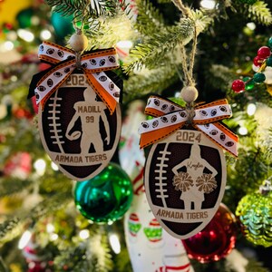 May include: Two wooden football-shaped Christmas ornaments. One features a football player silhouette, the other a cheerleader. Both have the text "Armada Tigers" and the year. Each ornament is adorned with a ribbon.