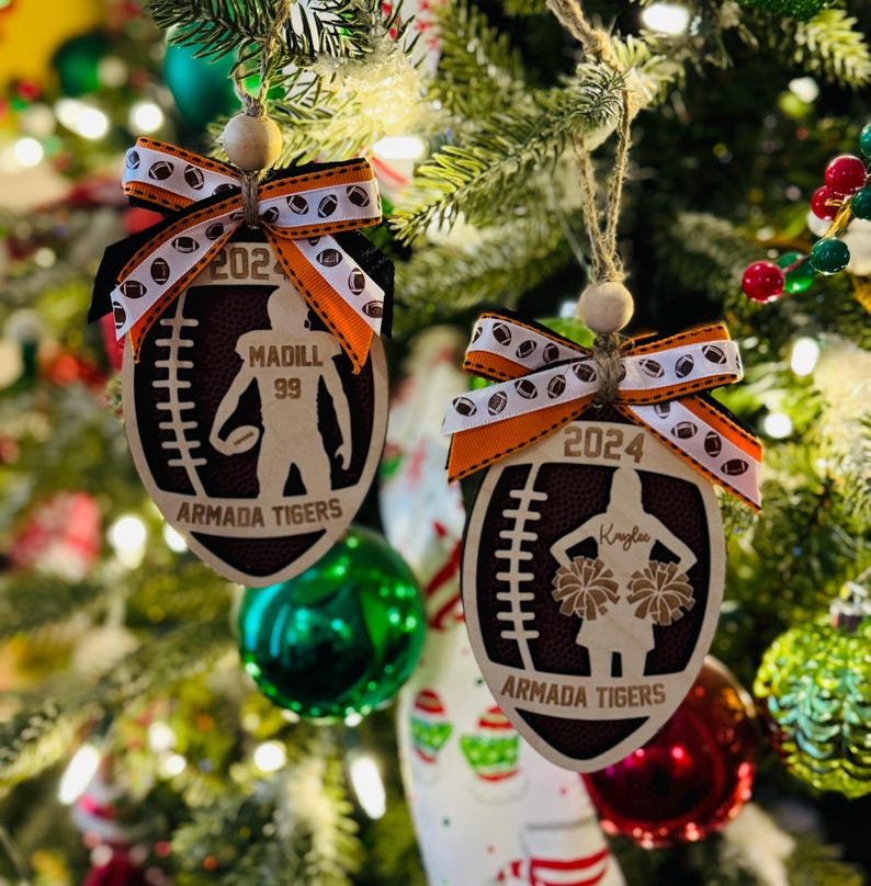 May include: Two wooden football-shaped Christmas ornaments. One features a football player silhouette, the other a cheerleader. Both have the text "Armada Tigers" and the year. Each ornament is adorned with a ribbon.