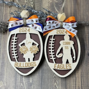 May include: Two wooden football-shaped ornaments. One features a cheerleader silhouette with the name "Eva" and the word "Bulldogs". The other has a football player silhouette with the name "Cobb" and the number "55", with the word "Eagles". Both have "2024" and are adorned with ribbons.