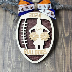 May include: Wooden football-shaped ornament with a brown football texture, a cheerleader silhouette, and the text "2024" and "BULLDOGS". It has a blue and orange ribbon with football graphics and a wooden bead.