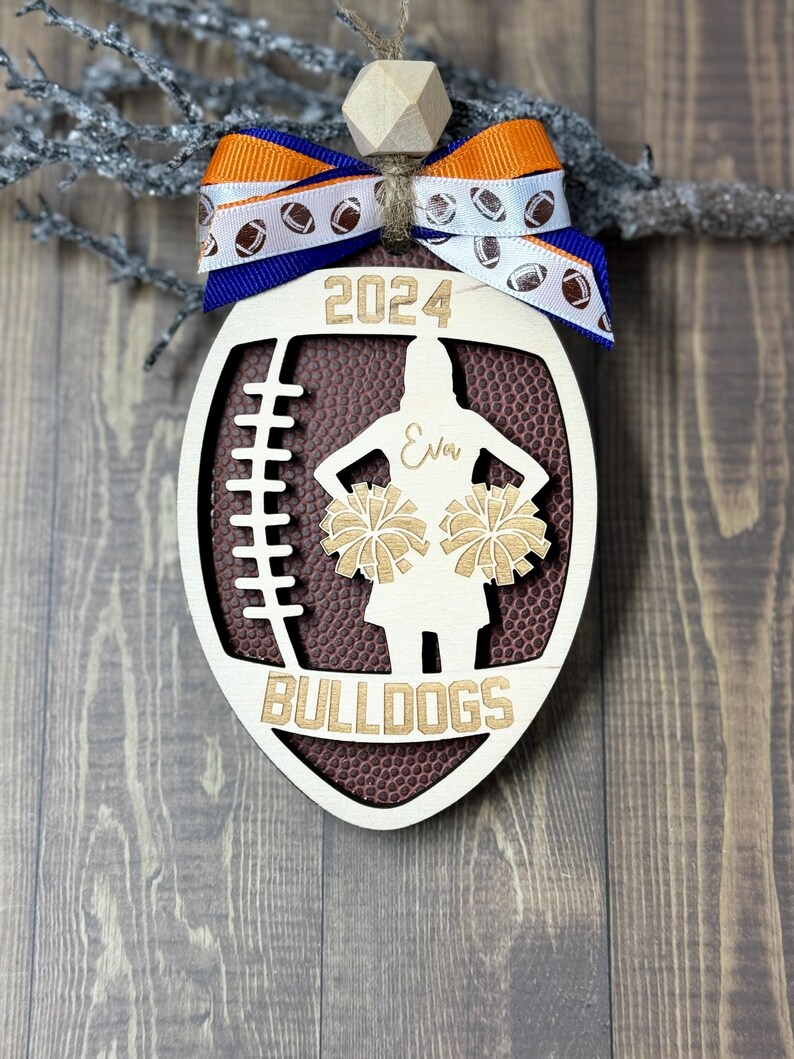 May include: Wooden football-shaped ornament with a brown football texture, a cheerleader silhouette, and the text "2024" and "BULLDOGS". It has a blue and orange ribbon with football graphics and a wooden bead.