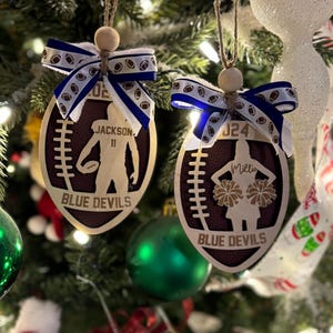 May include: Two wooden football-shaped Christmas ornaments. One features a football player silhouette, the name "Jackson" and the number "11". The other shows a cheerleader silhouette with the name "Millie" and the number "24". Both have "Blue Devils" text.