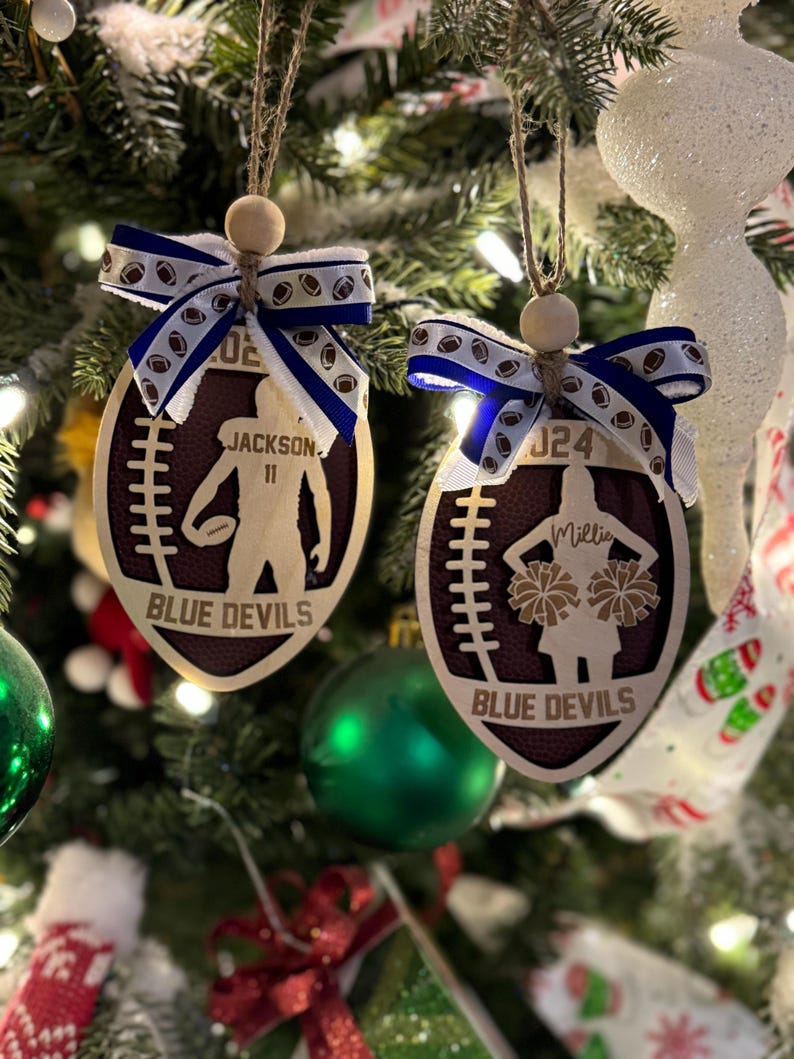 May include: Two wooden football-shaped Christmas ornaments. One features a football player silhouette, the name "Jackson" and the number "11". The other shows a cheerleader silhouette with the name "Millie" and the number "24". Both have "Blue Devils" text.