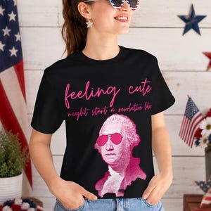 May include: A black t-shirt featuring a pink graphic of George Washington wearing pink sunglasses. The text in pink script reads, "feeling cute" above the text "might start a revolution later" in a smaller, simpler pink font.