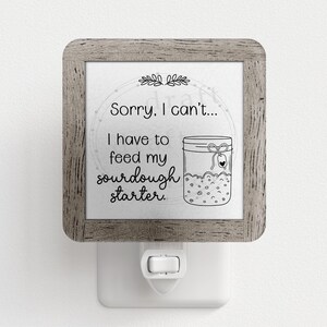 May include: A white night light with a wooden frame and a black and white illustration of a jar of sourdough starter. The text on the night light reads "Sorry, I can't... I have to feed my sourdough starter."