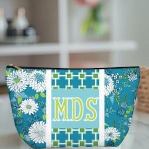 Könnte beinhalten: Türkisfarbene Kosmetiktasche mit floralem Muster und den Initialen "MDS" in Grün. Die Tasche hat einen Reißverschluss und ein geometrisches Design in der Mitte. Weiße und türkisfarbene Blumen sind über die Oberfläche der Tasche verstreut.