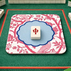Könnte beinhalten: Ein Mahjong-Spiel im Gange, mit elfenbeinfarbenen Spielsteinen und einem zentralen Spielstein mit einem roten chinesischen Schriftzeichen. Das Spiel befindet sich auf einer grünen Filzoberfläche mit einer floral gemusterten Matte in Rosa und Weiß.