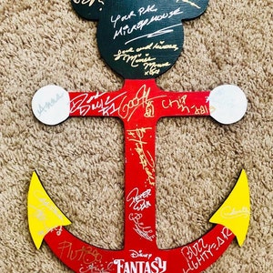 May include: A red wooden anchor with yellow tips, decorated with signatures from Disney characters. The anchor is shaped like Mickey Mouse's head with black ears. The text "Disney FANTASY 2019" is written on the anchor.