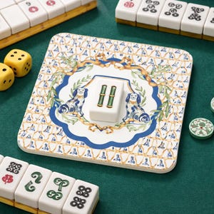 Könnte beinhalten: Ein quadratischer, dekorativer Mahjong-Steinehalter mit blau-weißem Blumenmuster, das zwei Hundeillustrationen zeigt. Ein weißer Stein mit grünen Bambussymbolen liegt auf dem Halter. Gelbe Würfel und andere Mahjong-Steine umgeben den Halter auf einer grünen Oberfläche.