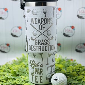 May include: White stainless steel tumbler with a golf-themed design. It features the text "Weapons of Grass Destruction" and "Let's Par Tee". A golf ball and tee are in the foreground.