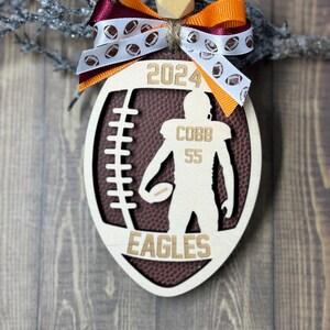 May include: Wooden football-shaped ornament with a football design, the year 2024, and the name "COBB 55" with "EAGLES" at the bottom. The ornament is decorated with a bow in maroon and orange.