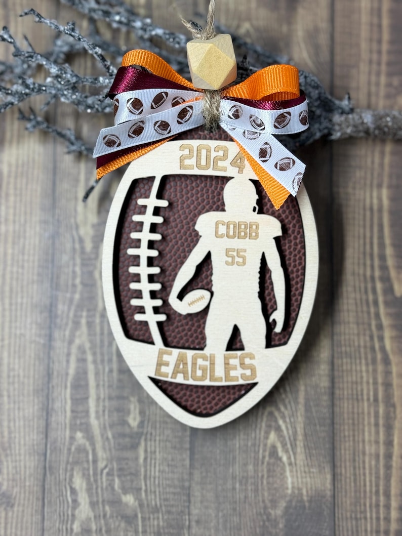 May include: Wooden football-shaped ornament with a football design, the year 2024, and the name "COBB 55" with "EAGLES" at the bottom. The ornament is decorated with a bow in maroon and orange.