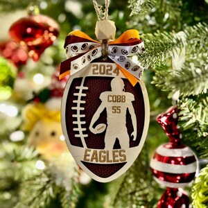 May include: Wooden football-shaped Christmas ornament with the text "2024", a football player silhouette, and "COBB 55 EAGLES". The ornament has a brown and white ribbon bow and is hanging on a Christmas tree.
