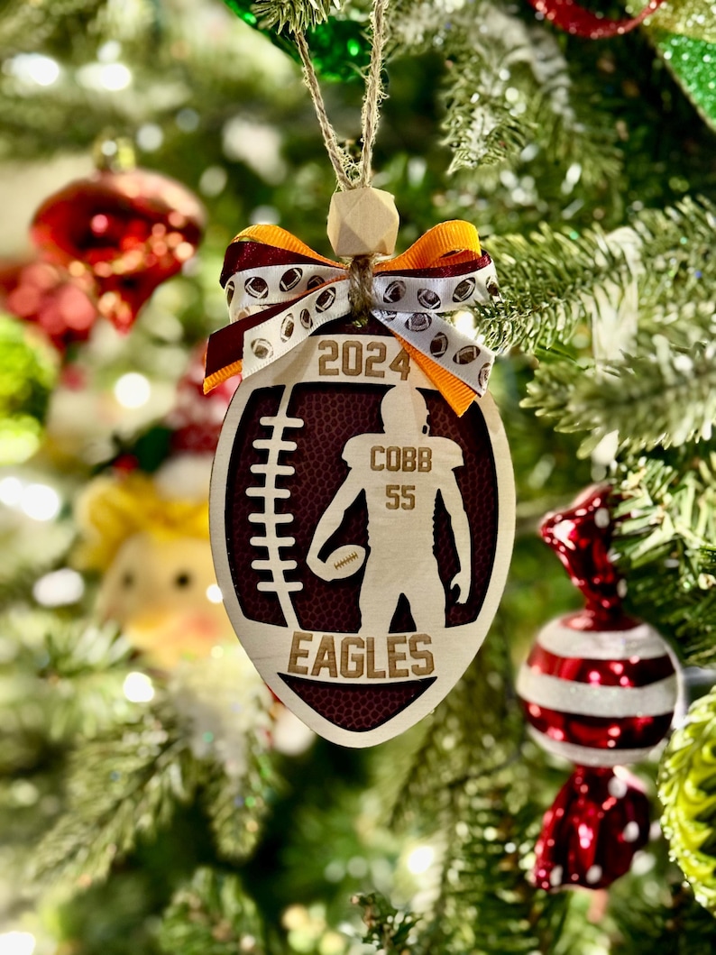 May include: Wooden football-shaped Christmas ornament with the text "2024", a football player silhouette, and "COBB 55 EAGLES". The ornament has a brown and white ribbon bow and is hanging on a Christmas tree.