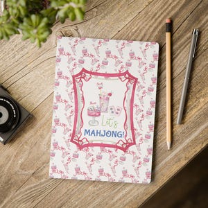 Op de afbeelding: Een notitieboek met een roze en wit patroon van taarten, strikken en cocktails. Het notitieboek heeft een roze kader met de tekst "Let's Mahjong!" en illustraties van cocktails en dobbelstenen.