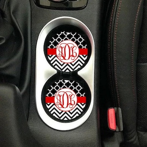 May include: Two black and white car coasters with a red stripe and a monogram in the center. The coasters are in a car cup holder.